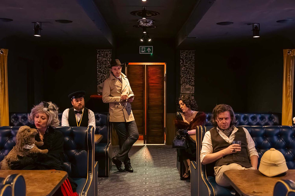 Passengers in vintage attire sit in a dimly lit train-style lounge, one man standing by the door under an exit sign.