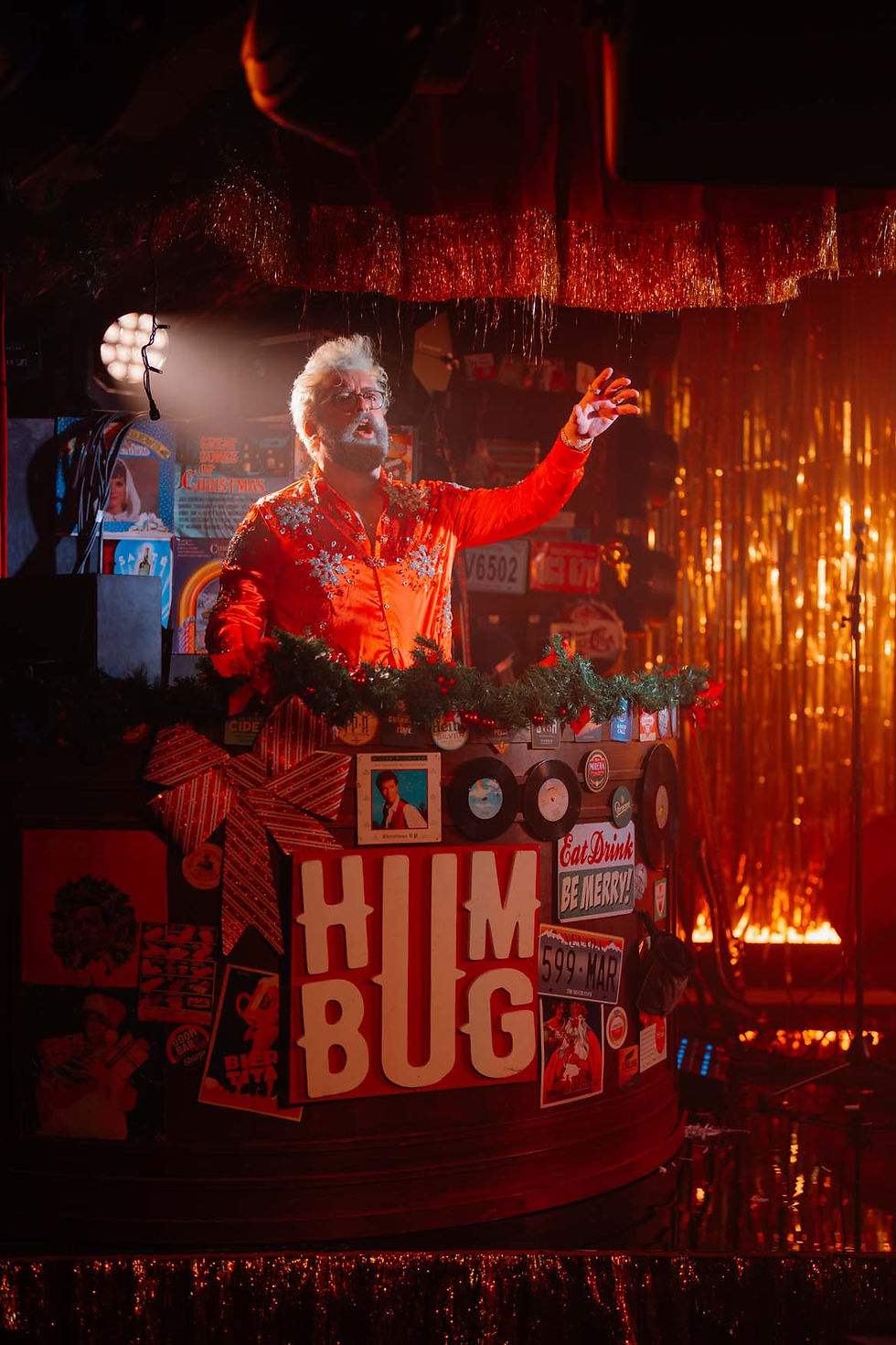 Person in red shirt stands in a festive setting with gold tinsel, gesturing over a decorated counter with "HUMBUG" sign and vintage decor.