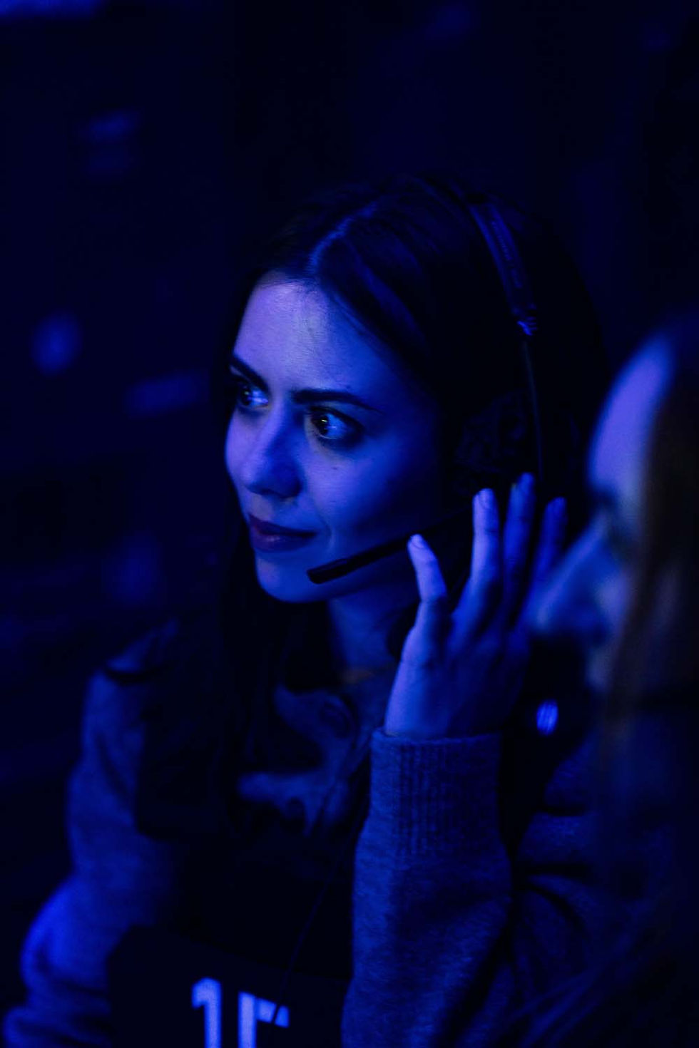 Woman with headset in dim blue light, focused expression. She's touching the headset, possibly communicating. Dark, blurred background.