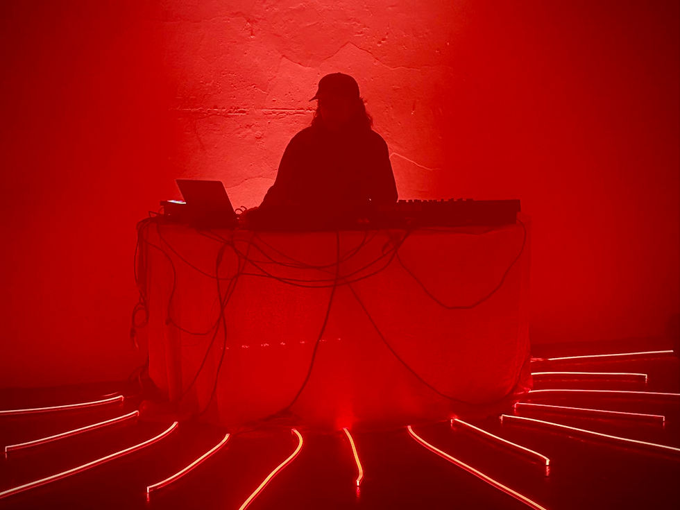 Silhouetted DJ with cap at a booth, surrounded by red lighting and cables. The room glows with an intense red ambiance.