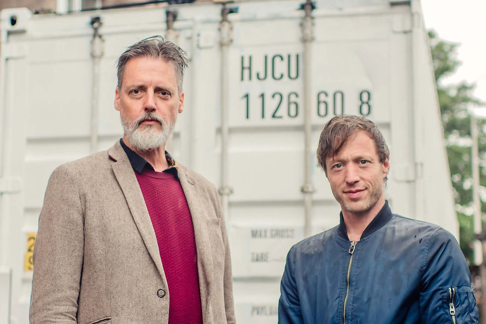 Two men stand in front of a shipping container. One wears a beige blazer, the other a navy jacket.