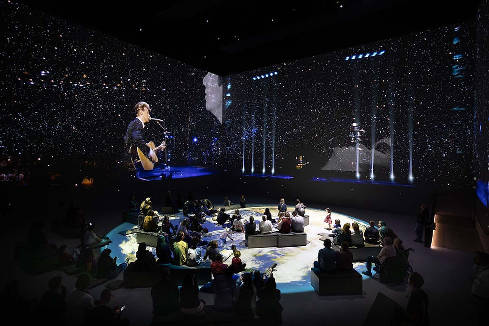 People seated in a dark room watch a large projection of a musician under a starry night sky, with Earth imagery on the floor.