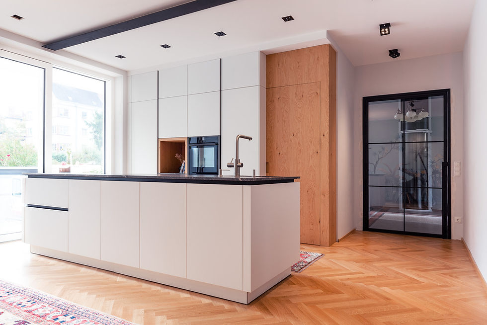 Eine moderne Küche mit einer großen Kücheninsel in weißer Hochglanzoptik und einer dunklen Arbeitsplatte. Die Insel hat klare, schlichte Fronten ohne sichtbare Griffe. Im Hintergrund sind hohe, weiße Einbauschränke mit einer Holzvertäfelung, die eine Nische mit einem eingebauten Backofen und einer silbernen Armatur an der Arbeitsplatte beherbergt. Links und rechts befinden sich große Fenster, die viel Tageslicht hereinlassen. Rechts neben den Schränken ist eine Glastür mit schwarzem Metallrahmen, die einen angrenzenden Raum abtrennt. Der Boden ist aus hellem Holz mit Fischgrätmuster verlegt. Der Raum wirkt hell, offen und elegant.