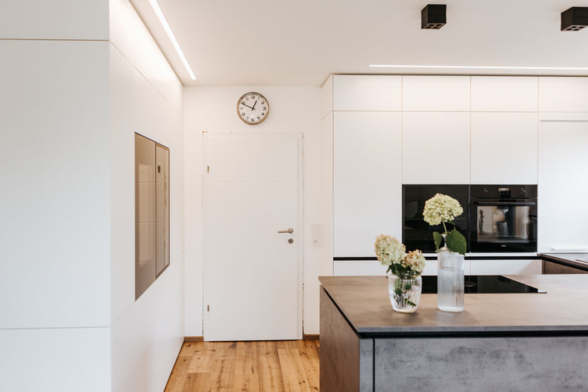 Ein heller Küchenbereich mit weißen Wänden und hellem Holzboden. An der linken Wand befinden sich zwei graue Bedienfelder, möglicherweise für Haustechnik oder Heizung. Rechts davon ist eine weiße Tür mit silbernem Griff. Über der Tür hängt eine runde Wanduhr mit weißem Zifferblatt und schwarzen Zahlen. Rechts ist ein Teil einer Kücheninsel mit dunklen Fronten zu sehen, auf der sich eine Glasvase mit weißen Blumen und eine dekorative Flasche befinden. Im Hintergrund sind weiße Einbauschränke mit integrierten schwarzen Küchengeräten erkennbar. Der Raum wirkt modern, sauber und einladend.