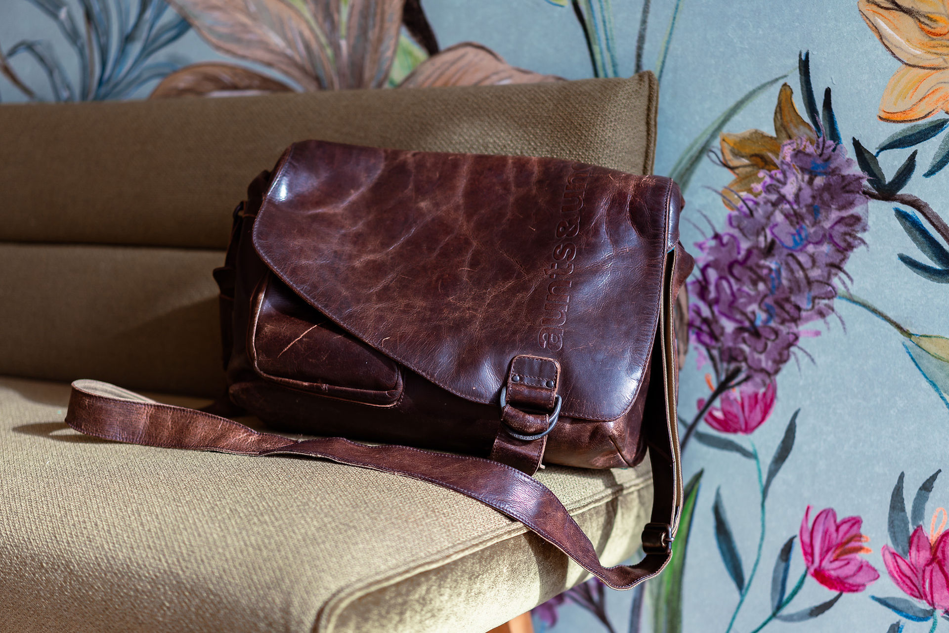 Eine braune Leder-Umhängetasche liegt auf einer braun-grünlichen Sitzbank. Die Tasche hat eine Klappe mit einer Schnalle und einen langen, verstellbaren Trageriemen. Im Hintergrund ist eine bunte Tapete mit floralen Mustern in verschiedenen Farben zu sehen. Neben der Sitzbank steht ein Strauß lila Blumen.