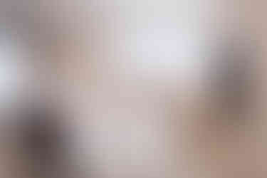 Moderne, minimalistische Küche in hellen Tönen mit großer weißer Kücheninsel im Vordergrund; darauf steht ein kleiner Obstkorb. Rechts sind hohe Einbauschränke mit Backofen, hinten Spüle unter einem breiten Fenster.