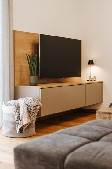 Wohnbereich mit Wandverbau aus Holz und hellem Lowboard, darauf Fernseher, Pflanze und Tischleuchte, im Vordergrund dunkelgraue Couch und geflochtener Korb mit Decke