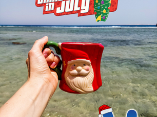 A hand holds a festive red Santa Claus mug against the backdrop of a crystal-clear turquoise ocean on a sunny day. The playful holiday mug contrasts with the tropical beach setting, capturing the fun spirit of Christmas in July with calm waters, rocky shoreline, and a bright blue sky.