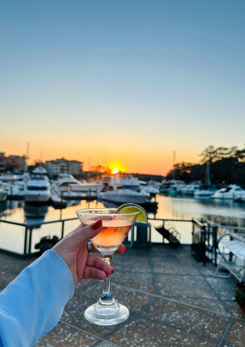 a hand holding up a cocktail with the sun setting over the marina in the background