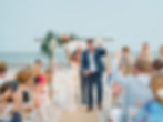 Bride and groom celebrating on a beach under a floral arch. Guests cheer and take photos. Bright, joyful atmosphere by the sea.