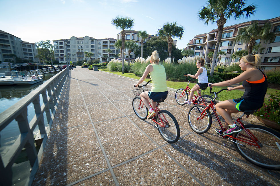 Three women ride red bikes along a sunny marina path beside palm trees and resort condos.