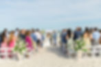Beach wedding ceremony with a couple under a floral arch, surrounded by guests seated on white chairs. Ocean and blue sky in the background.