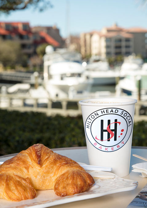 closeup of a croissant and coffee cup with the Hilton Head Social logo and marina in the background
