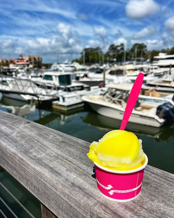 Bright yellow Lemon gelato from Frosty's in a pink cup with a spoon sits on a railing at Shelter Cove marina. Boats and a blue sky with clouds are in the background.