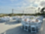 Outdoor event setup with round tables and white chairs, decorated with flowers and tableware. String lights above; grassy dunes in background.