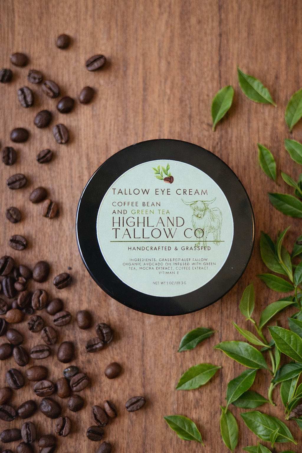 black jar with a green label on a wooden back ground with coffee beans and tea leaves. the label states tallow eye cream