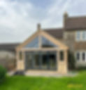 Modern house extension with large glass windows and wooden frame, blending contemporary design with traditional stone architecture. Green lawn in front and Selfix Builders logo.