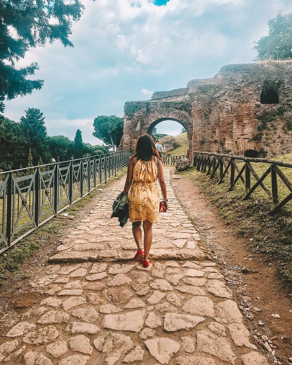 Woman in a yellow dress walking along the ancient stone path of Palatine Hill towards a brick arch, photographed by shiniestvoyager. By a colosseum audio guide review 2025