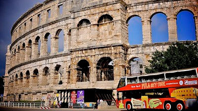 Open‑top tour bus parked beside a massive stone amphitheatre with sunlit limestone Colosseum framed by a deep blue sky.