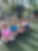Young children sitting together on a playground surface outdoors, smiling and looking at the camera during a Temple Sinai early childhood program, representing the Early and After Care experience for children ages 18 months to 5 years old.