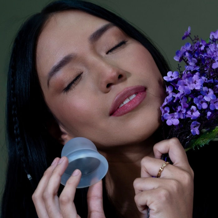 Mujer Latina con ojos cerrados y flores lilas en una mano y un disco de menstruacion en su otra mano