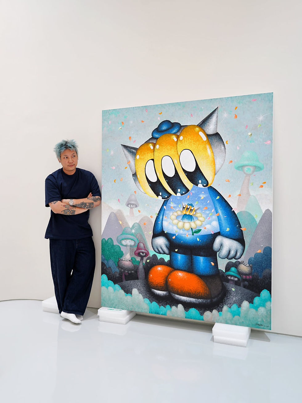 Man with blue hair stands beside a colorful surreal painting in a white gallery, arms crossed, calmly posing.