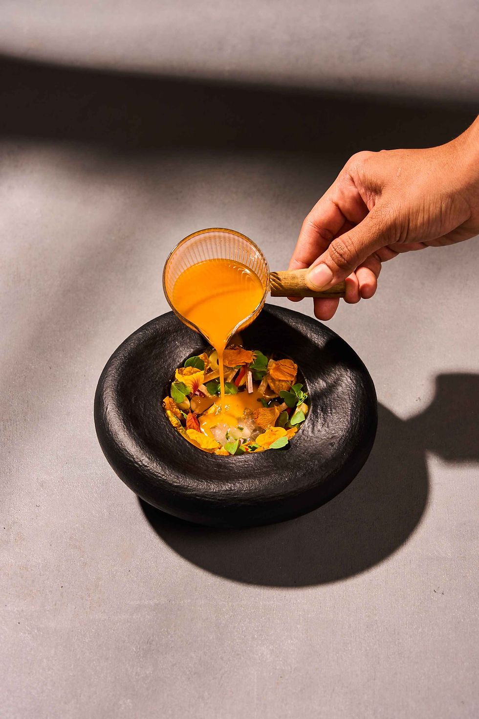 A hand pours orange sauce from a small cup onto a dish with flowers and herbs in a black bowl on a gray surface. Bright and artistic.