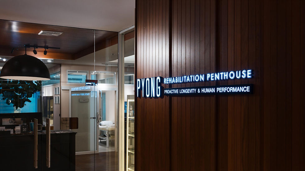 Neon sign on a wood wall reads PYONG Rehabilitation Penthouse, Proactive Longevity & Human Performance in a sleek clinic lobby.