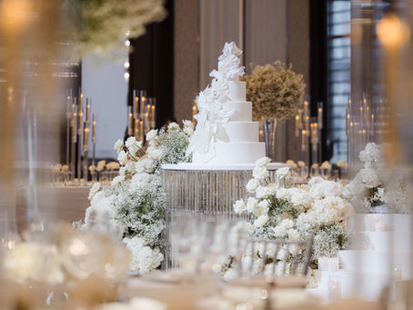 Elegant white wedding cake with floral decorations and candles on a table surrounded by white flowers in a luxurious, soft-lit setting.
