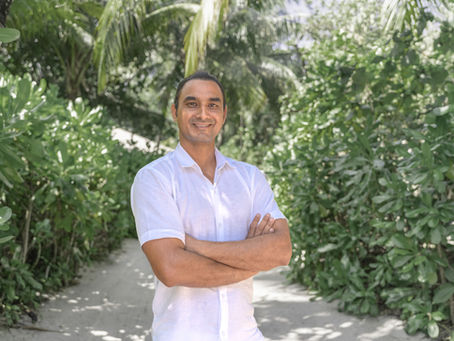 Smiling man in a white shirt stands with arms crossed on a sandy path amid lush tropical greenery.