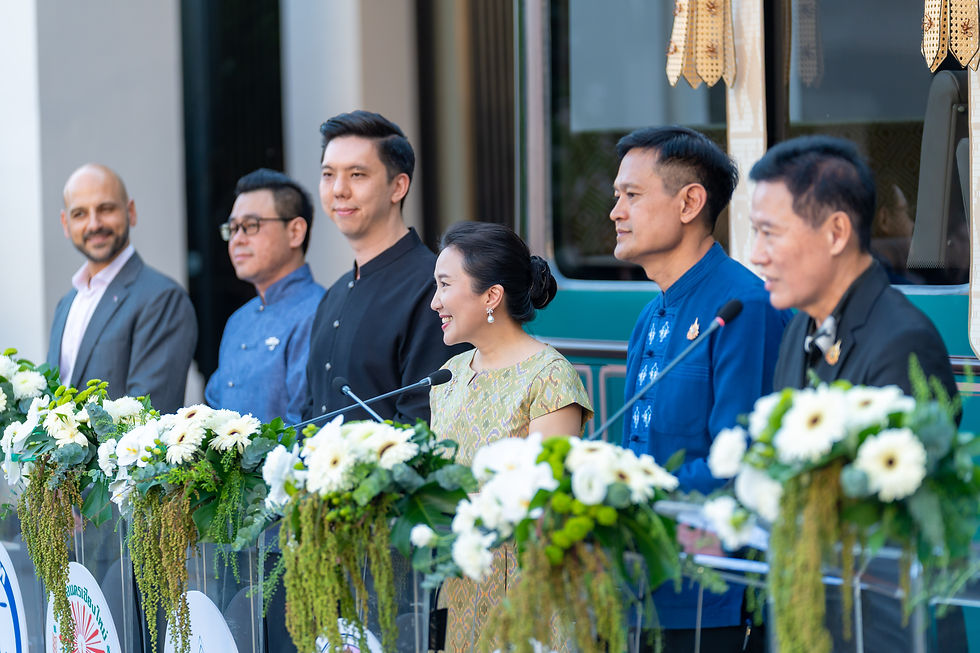 Five officials stand behind flower-lined podiums at an outdoor ceremony, smiling and speaking into microphones.