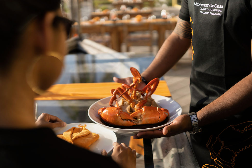 Waiter in a Ministry of Crab shirt serves a plate of crab to a diner at an outdoor table in warm sunlight.