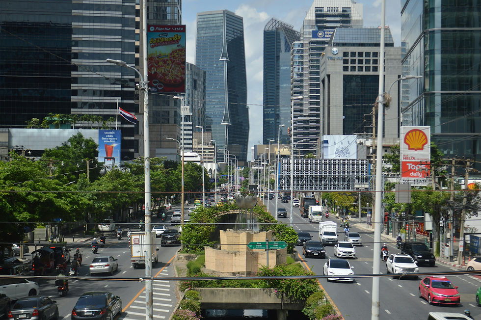 Busy Bangkok boulevard with cars and motorbikes between tall glass towers, Shell and Pizza Hut signs, under a bright sky.