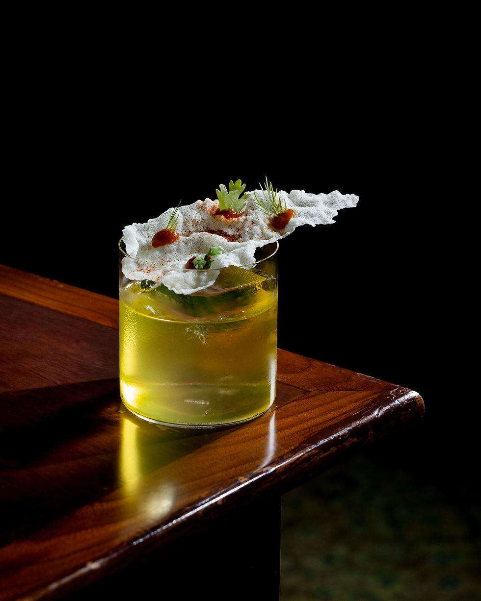 Yellow cocktail in a low glass on a wooden table, topped with airy white garnish, herbs, and red dots against a black background