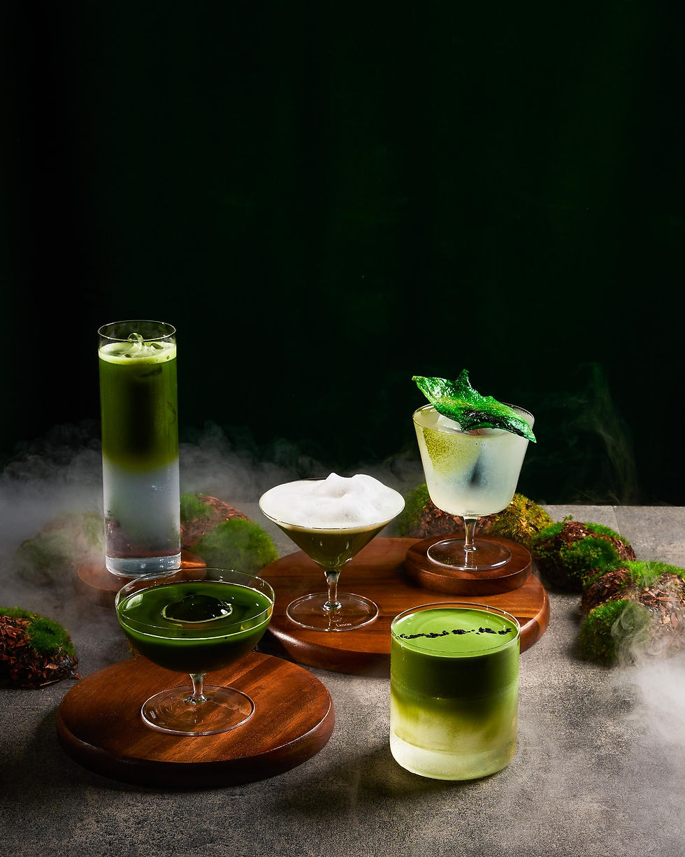 Five green cocktails on wooden stands amid fog, one topped with foam and another with a leaf, against a dark background.