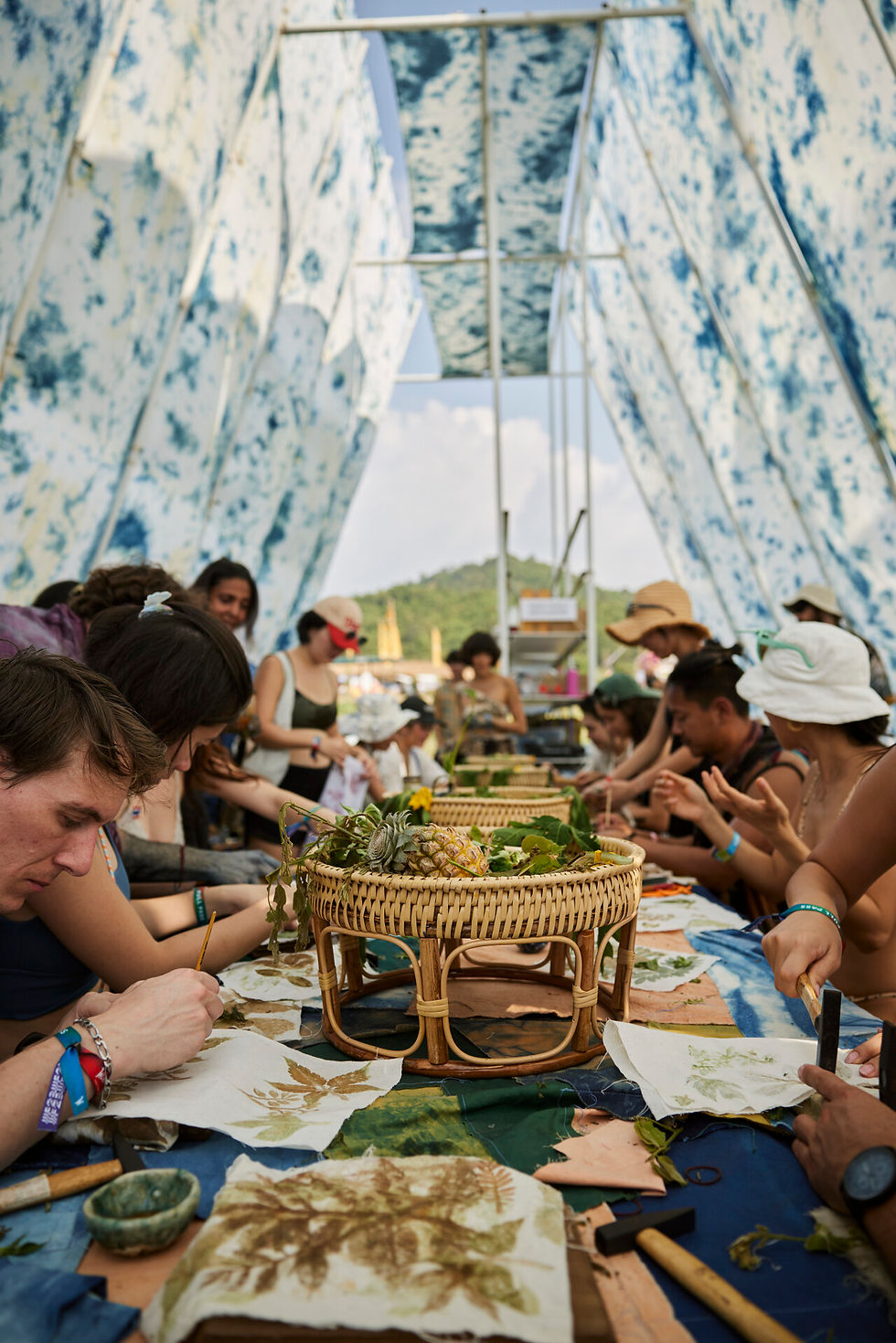 People make leaf prints at a long table under a blue-white canopy, with baskets and tropical hills in the background.