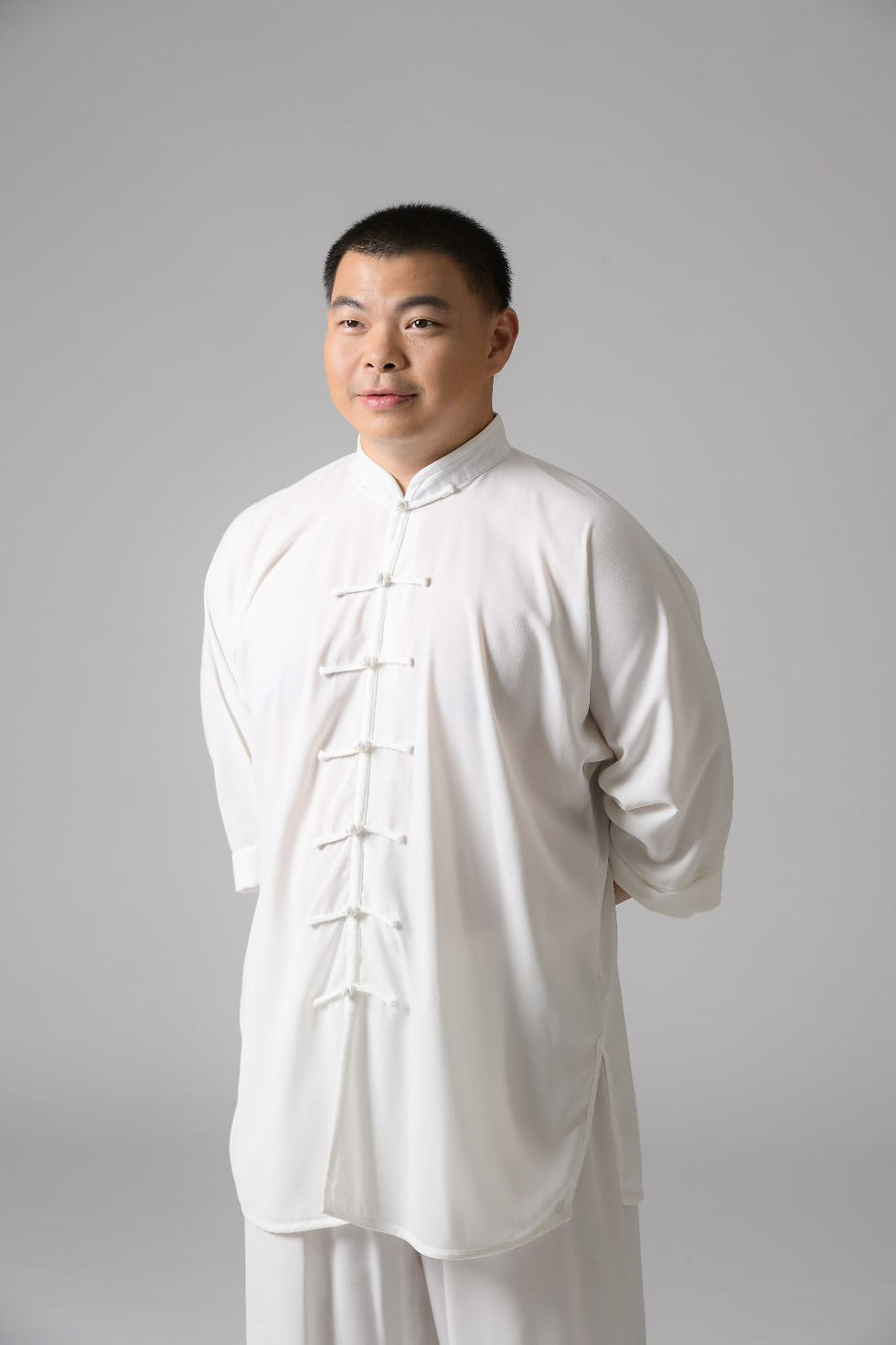 A person stands smiling in a white traditional outfit against a plain gray background. The mood is calm and serene.
