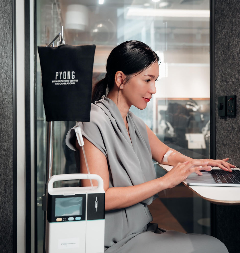 Woman in gray dress typing on a laptop beside an IV drip labeled PYONG in a bright clinic office, focused and calm