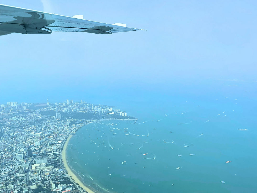 Aerial view from an airplane wing over a coastal city, curved bay, and boats on calm blue water under a hazy sky.