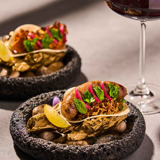 Goan sausage taco served on a dark stone plate with herbs, pickled onions, and lime, paired with a glass of red wine in an elegant fine dining setting.