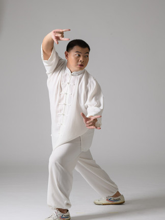 A person in white martial arts attire strikes a dynamic pose, smiling slightly in a plain, light-colored studio setting.
