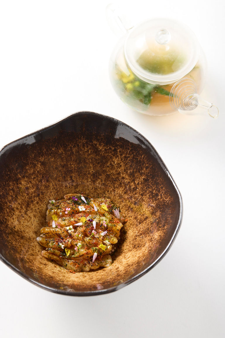 Top-down of a dark bowl with a small gourmet dish topped with flower petals, beside a glass teapot on a white background.