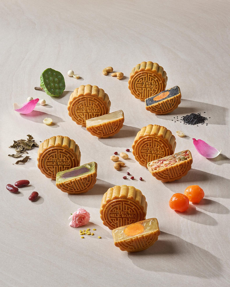 Assorted mooncakes embossed THE SILK ROAD, arranged with nuts, petals, and seeds on a beige studio background.