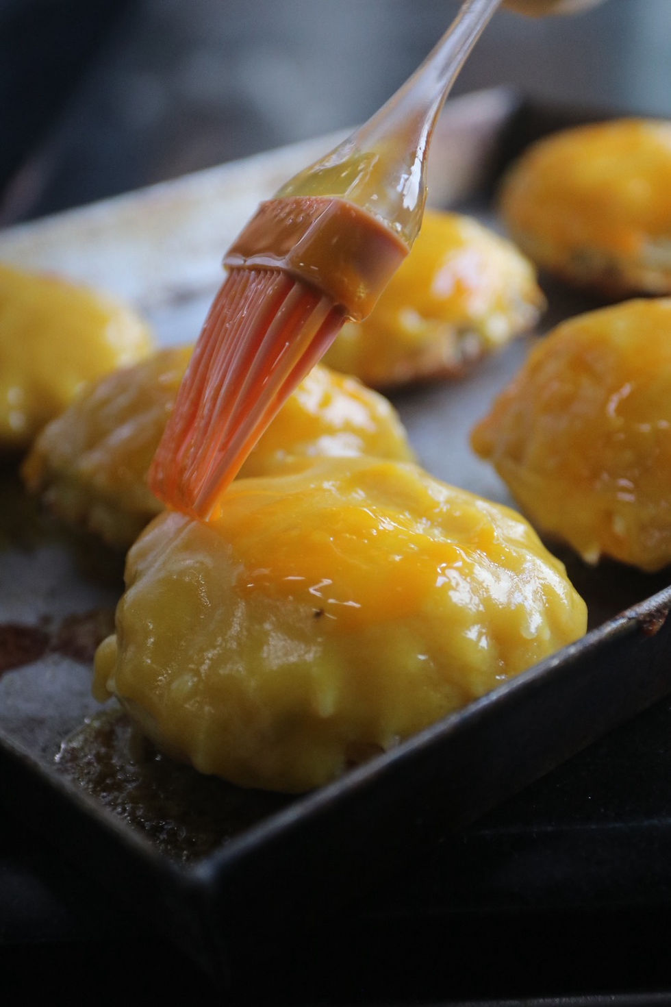Pastry buns on a baking tray being brushed with egg wash, glossy yellow tops in a close-up kitchen prep scene.