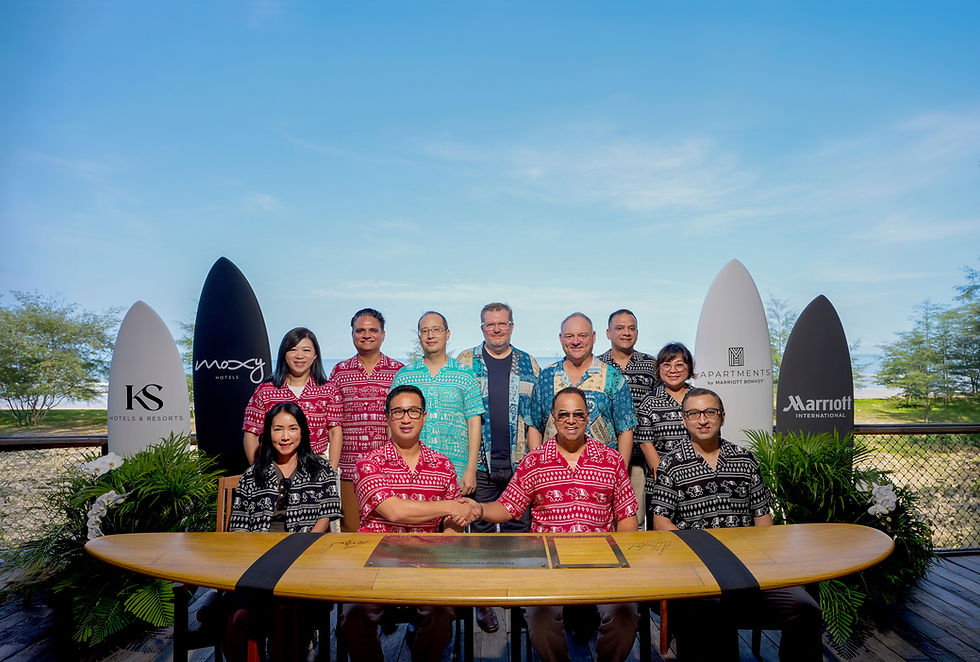 Group of men and women in colorful Hawaiian shirts pose outdoors by a surfboard display, shaking hands at a signing table.