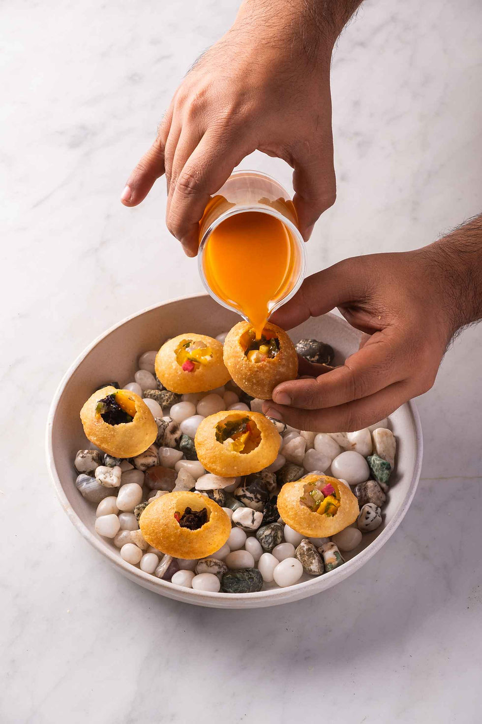Hands pour orange liquid into golgappas in a bowl of colorful pebbles on a marble surface, creating a vibrant and appetizing scene.