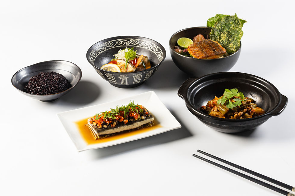 An assortment of dishes on a white background: black rice, fish, and vegetables in black bowls, and a plated fish with sauce and herbs.