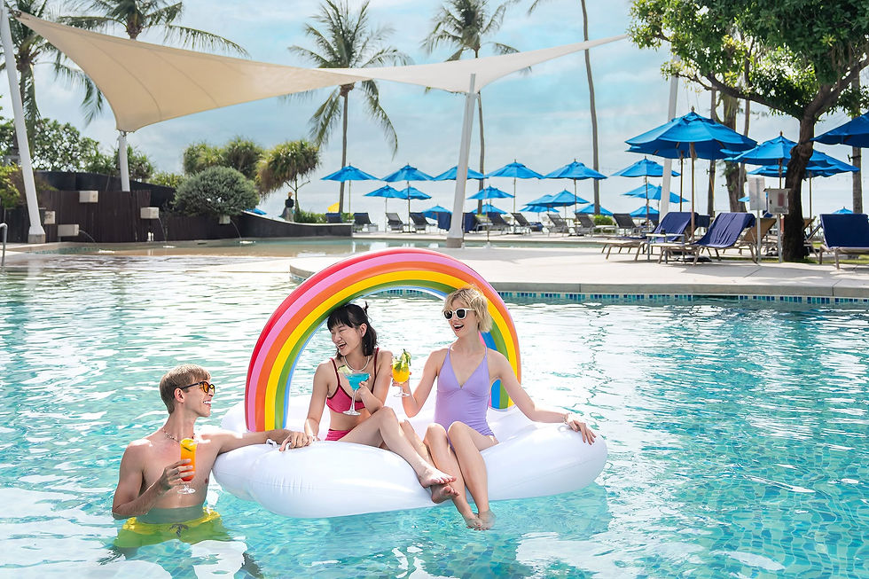 Three friends in a resort pool on a rainbow float, laughing with drinks under blue umbrellas and palm trees.