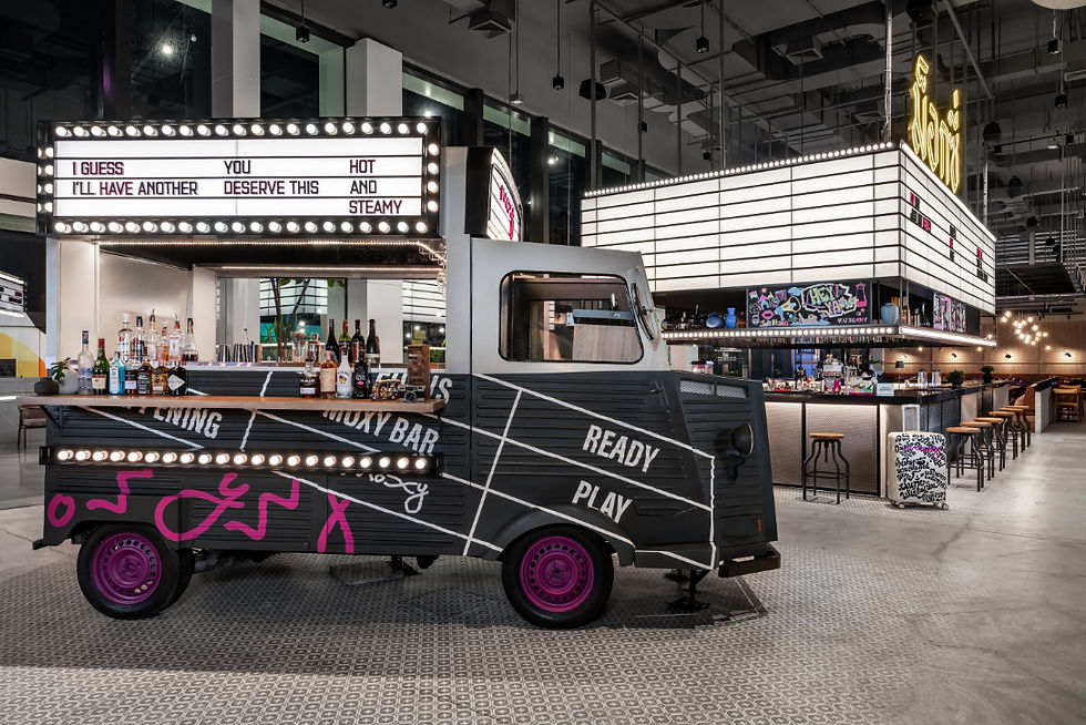 Truck-shaped bar in a modern venue, lit by marquee bulbs, with bottles and stools; signs read I GUESS I’LL HAVE ANOTHER and READY PLAY