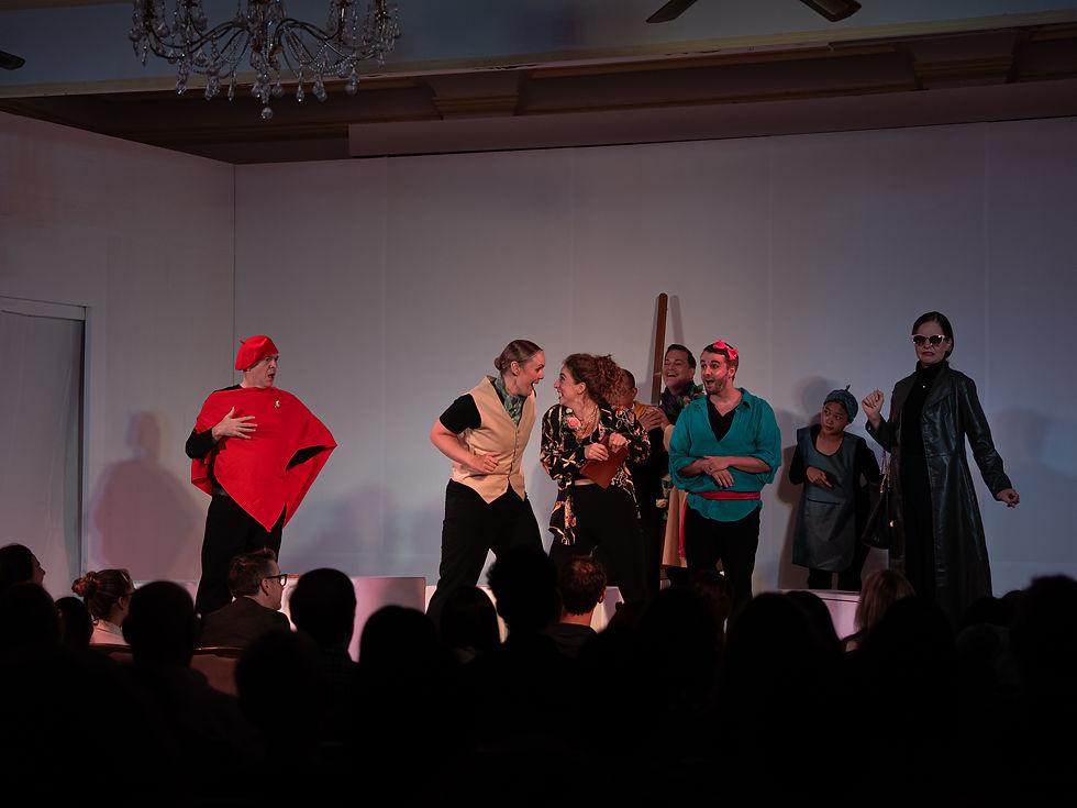 Actors in colorful costumes perform on a stage before a seated audience, laughing and gesturing under soft theater lighting.